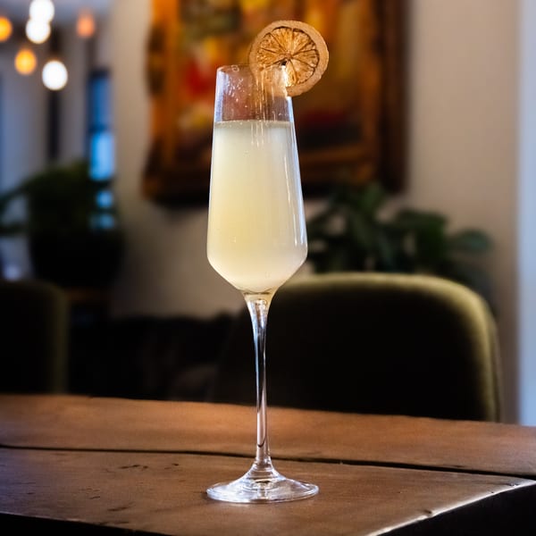 A tall glass of light-colored cocktail with a dried orange slice on a wooden table.