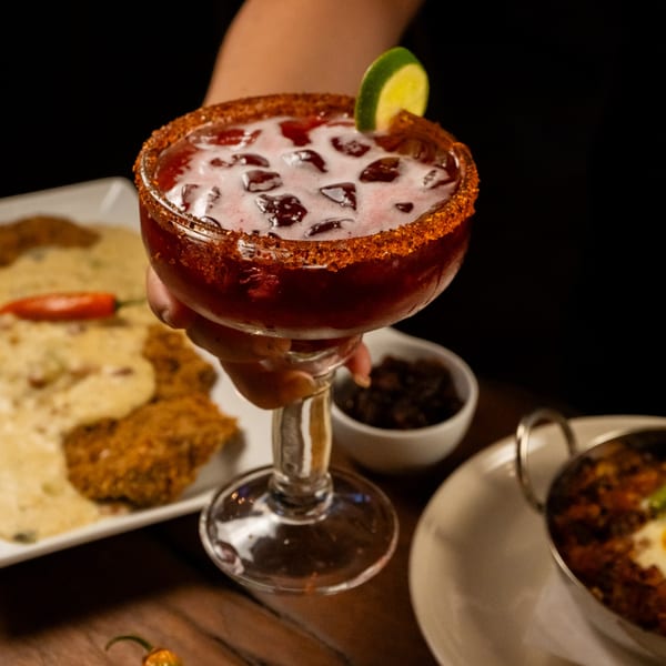 A hand holds a red cocktail with a chili-salt rim and lime, with food in the background.