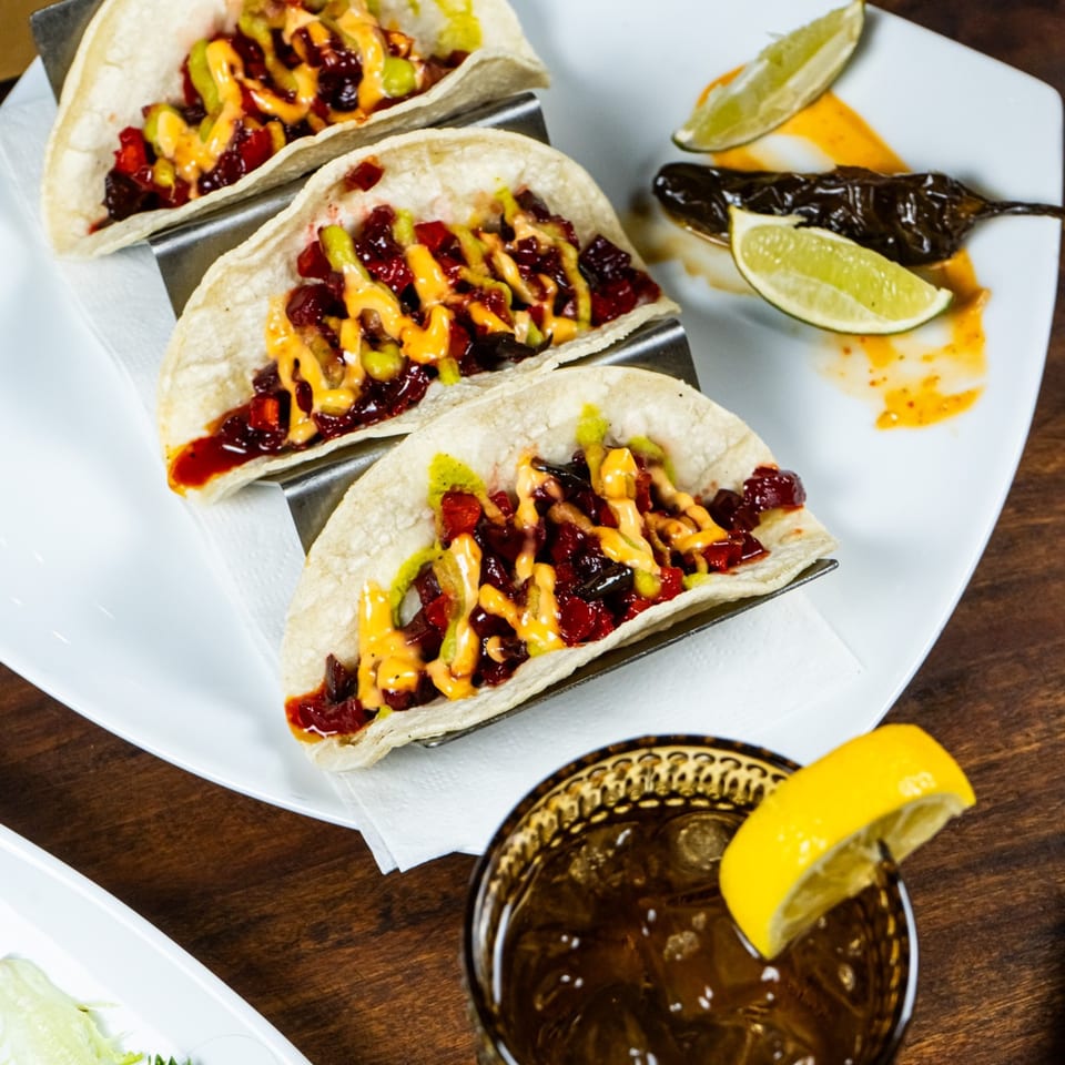 Three colorful tacos with red and green toppings, drizzled with sauce, next to a cocktail.